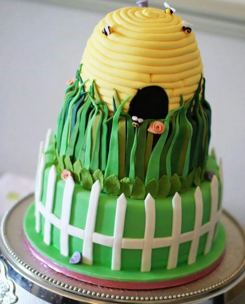 Garden-themed celebration cake with a yellow beehive top tier, sugar-paste bees, long grass and a white picket fence