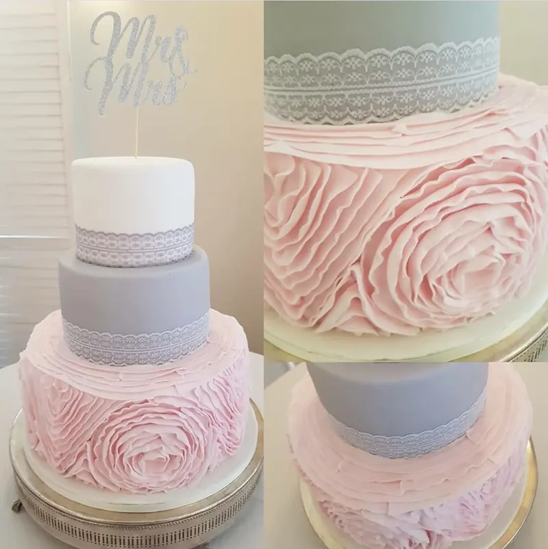 Three-tier wedding cake with a pink ruffled rose bottom tier, grey and white tiers trimmed with lace, and a glittery Mrs & Mrs topper